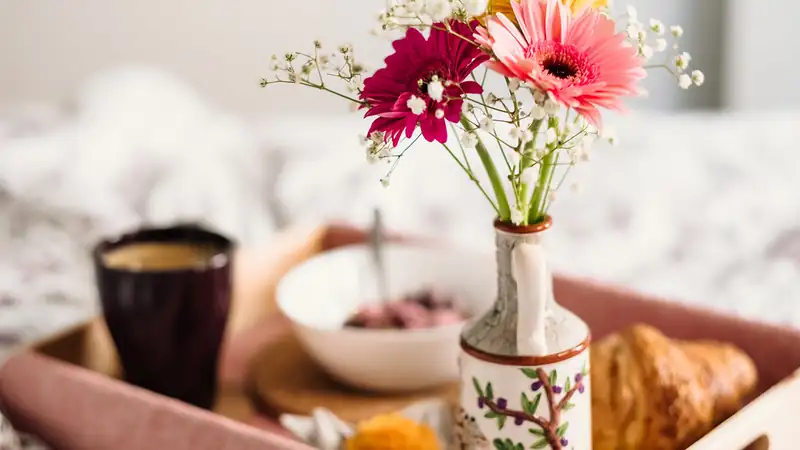 A vase of pink gerberas and baby's breath beside coffee and a croissant on a tray.