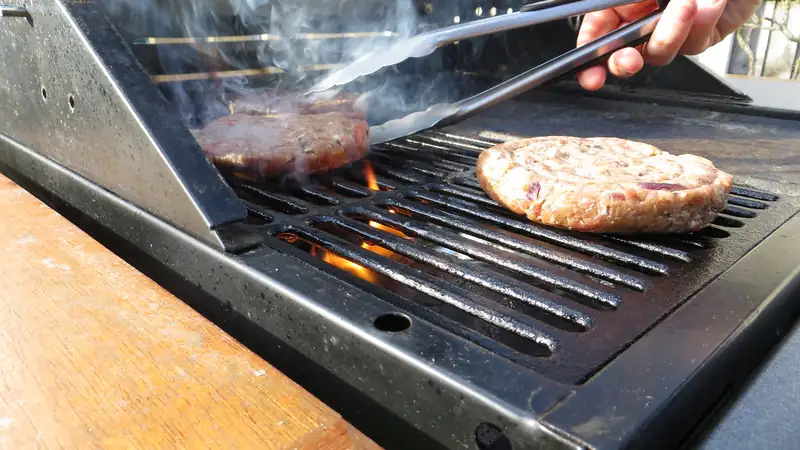 Tongs flipping burger patties on a smoky backyard grill.