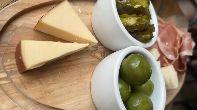 A wooden board with smoked gouda wedges, prosciutto, olives and pickles.