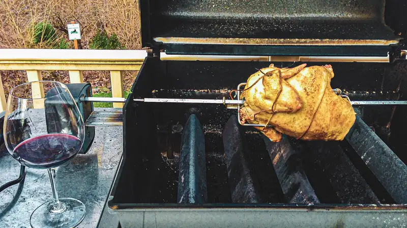 A whole chicken turning on a rotisserie spit in a grill beside a glass of red wine.