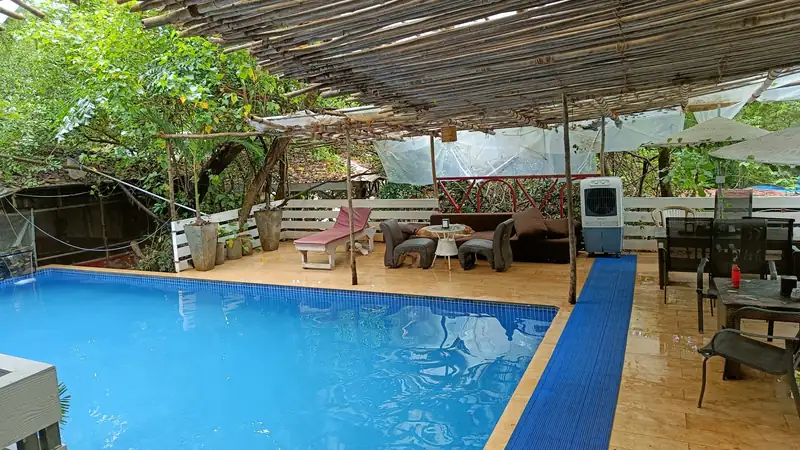 A swimming pool with blue deck mats beside a shaded patio with chairs.