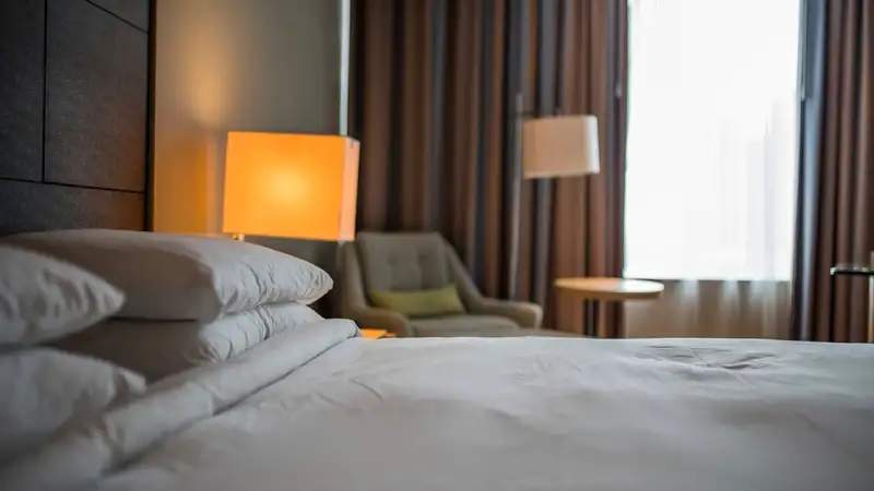 A made bed with plump white pillows beside a glowing lamp and an armchair by the window.
