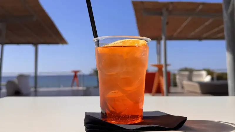 An orange cocktail with ice, an orange slice and a black straw on a sunny terrace table.