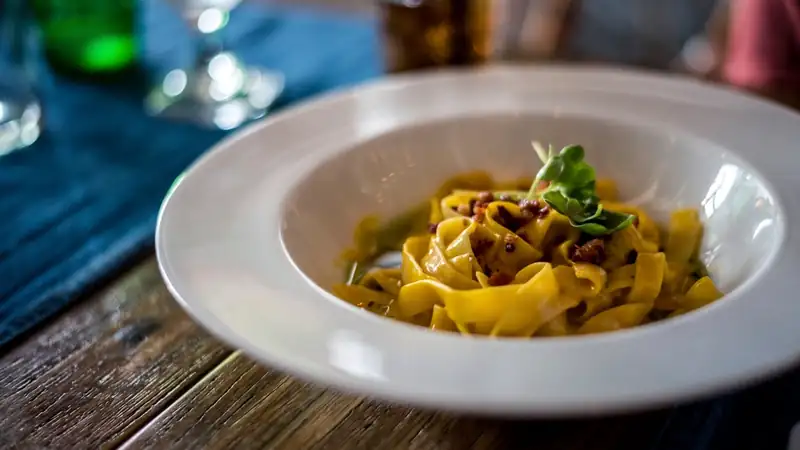 A wide white bowl of tagliatelle with meat ragù and herbs on a wooden table.